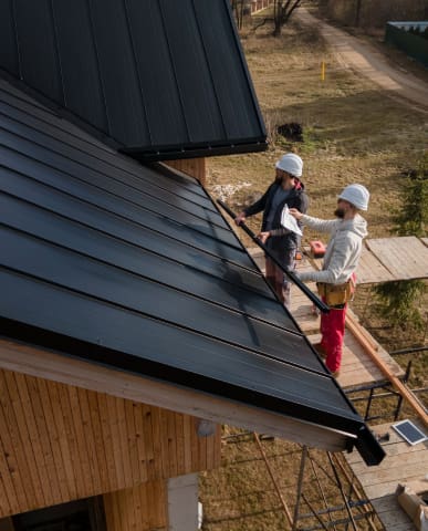 Two workers installing a black metal roof on a building.
